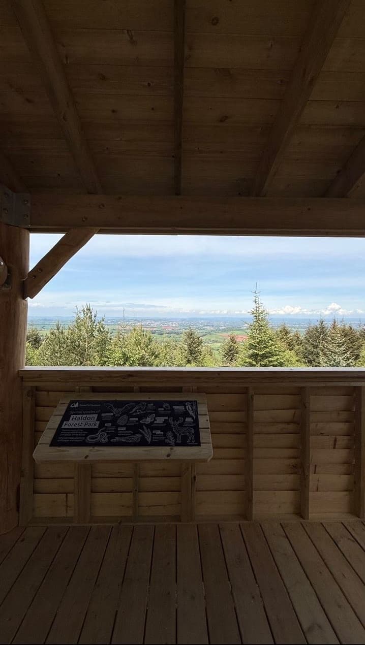 View from inside the shelter looking out over the Devon countryside, with a Forestry England wildlife information sign mounted on the wall