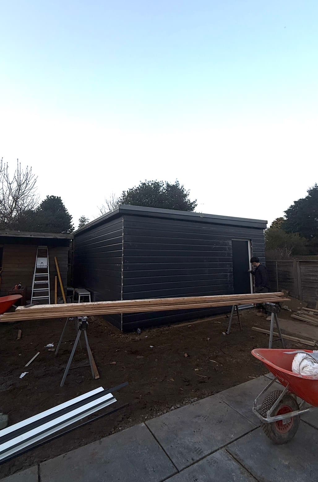 Black-clad, flat-roofed timber garden room nearing completion, with the door being fitted