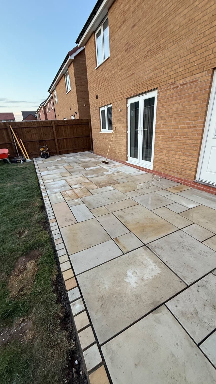 Patio nearing completion at a new-build brick house in evening light, with tools and a broom still on site