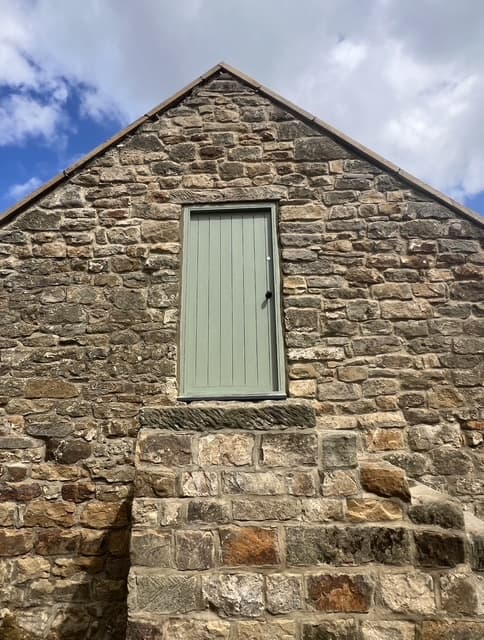 Close detail of the stone gable end and a matching sage-green door on the same rural outbuilding